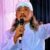 Male singer on stage, wearing a white robe and head wrap, holding a microphone and raising one arm.