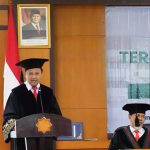 Man in an academic gown and cap delivers a speech at a podium during a formal ceremony with an indonesian flag a portrait and a banner in the background