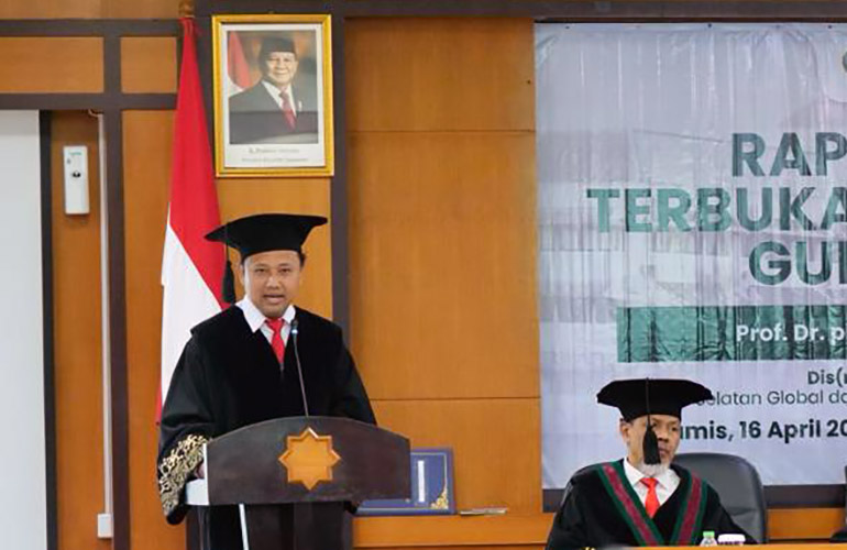 16dgubes catrawarta Man in an academic gown and cap delivers a speech at a podium during a formal ceremony with an indonesian flag a portrait and a banner in the background