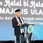 Older man in a black suit and white scarf speaks at a podium on a stage with a large backdrop of bold white text
