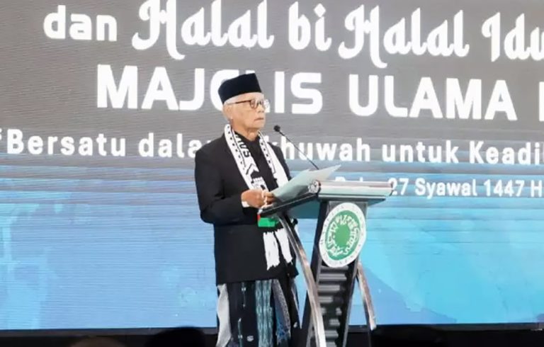 Older man in a black suit and white scarf speaks at a podium on a stage with a large backdrop of bold white text.