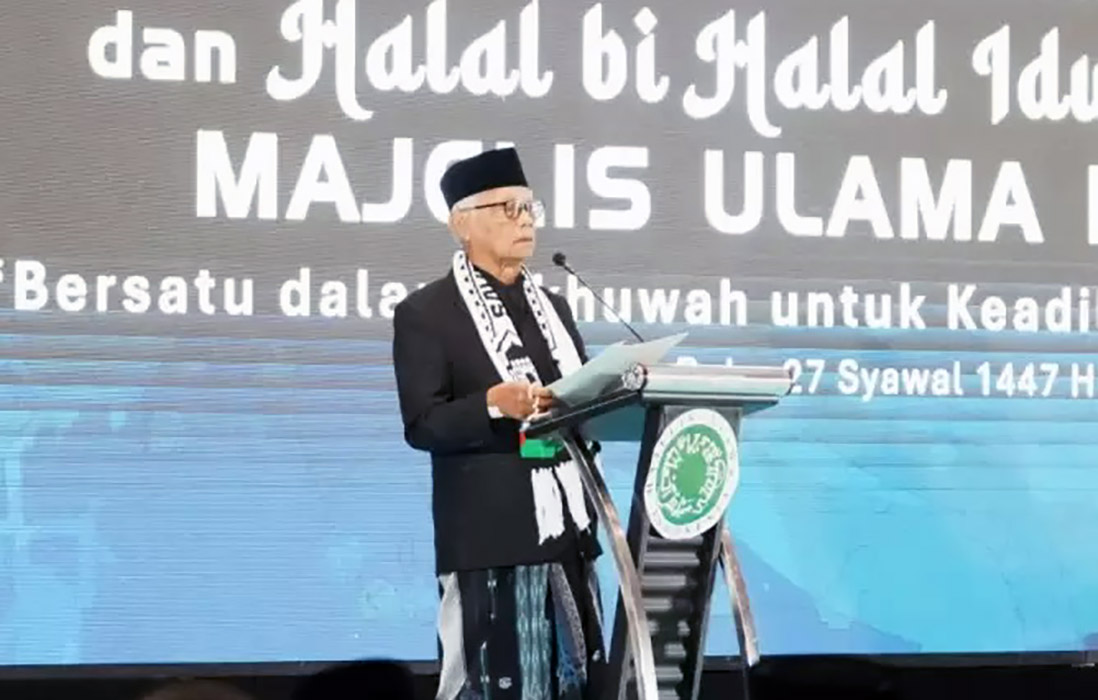 16dketum1 catrawarta Older man in a black suit and white scarf speaks at a podium on a stage with a large backdrop of bold white text