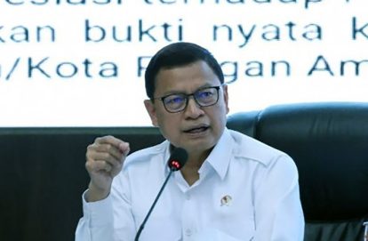 Man in a white shirt and glasses speaking into a microphone at a press conference or panel discussion in front of a text-filled backdrop.