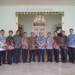 Twelve men in batik shirts stand in a line for a group photo in a decorated hall with ornate door frames and a chandelier overhead