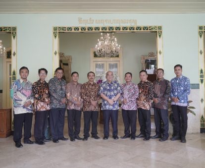 Twelve men in batik shirts stand in a line for a group photo in a decorated hall with ornate door frames and a chandelier overhead