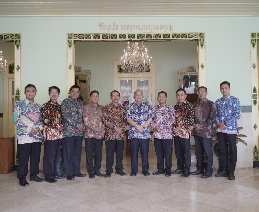 Twelve men in batik shirts stand in a line for a group photo in a decorated hall with ornate door frames and a chandelier overhead