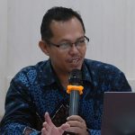 Man in glasses speaks into a handheld microphone while seated at a table with an asus laptop nearby
