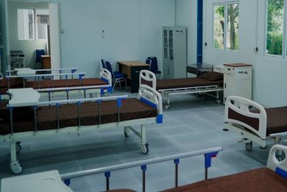 A ward with multiple hospital beds on wheels, blue railings, and brown mattresses arranged along light blue tiled floor.