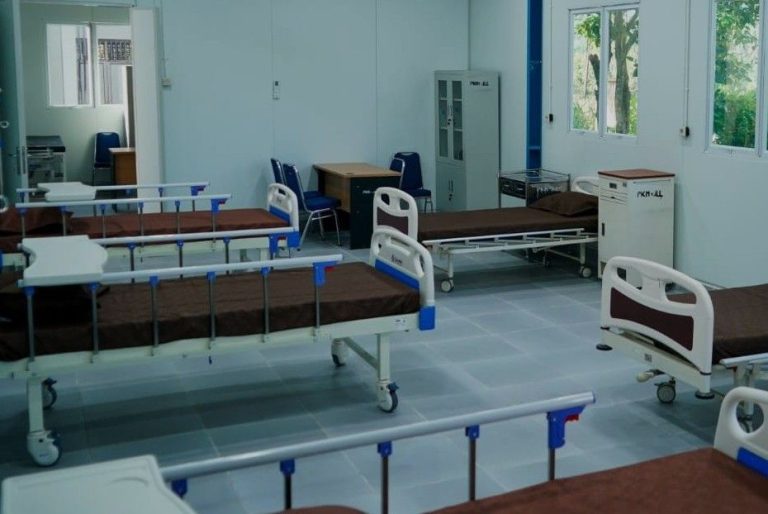 A ward with multiple hospital beds on wheels, blue railings, and brown mattresses arranged along light blue tiled floor.