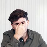 Man in a green button up shirt pinching his nose with eyes closed looking fatigued against a light striped background