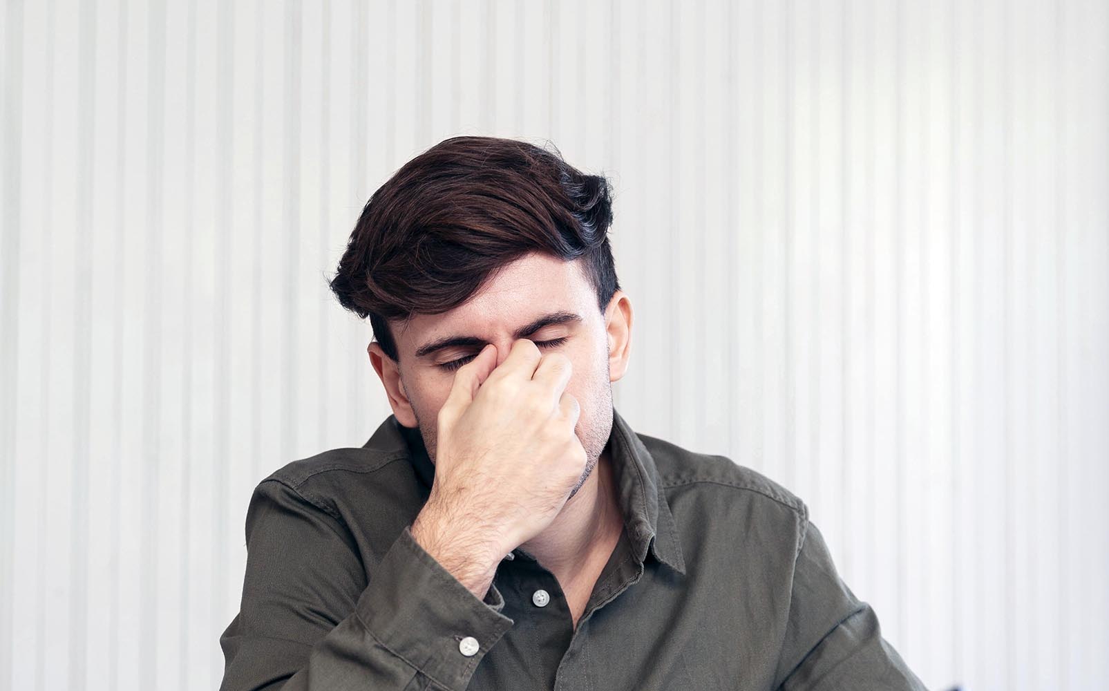 Man in a green button up shirt pinching his nose with eyes closed looking fatigued against a light striped background