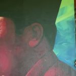 Mural of a laughing face on the left a bearded man peeks from behind a bright multicolored panel on the right