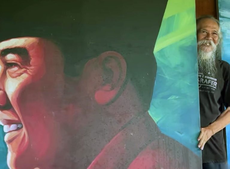 Mural of a laughing face on the left; a bearded man peeks from behind a bright, multicolored panel on the right.