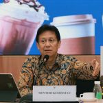 Man seated at a press conference microphone in front of a sign reading menteri kesehatan indonesian health minister with a tablet on his left and beverages on the table