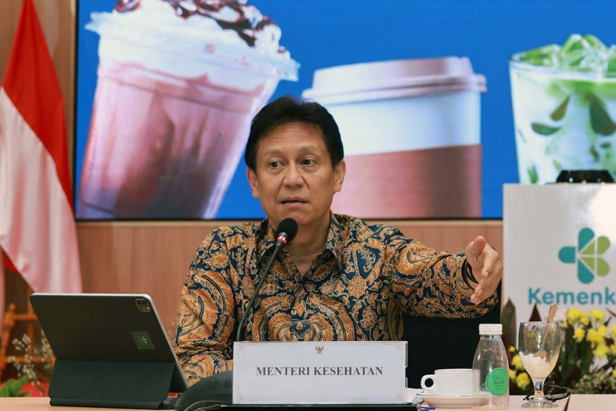 Man seated at a press conference microphone in front of a sign reading menteri kesehatan indonesian health minister with a tablet on his left and beverages on the table
