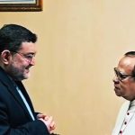 Two men in conversation one in a dark suit with hands clasped the other in white clerical attire with glasses nearby