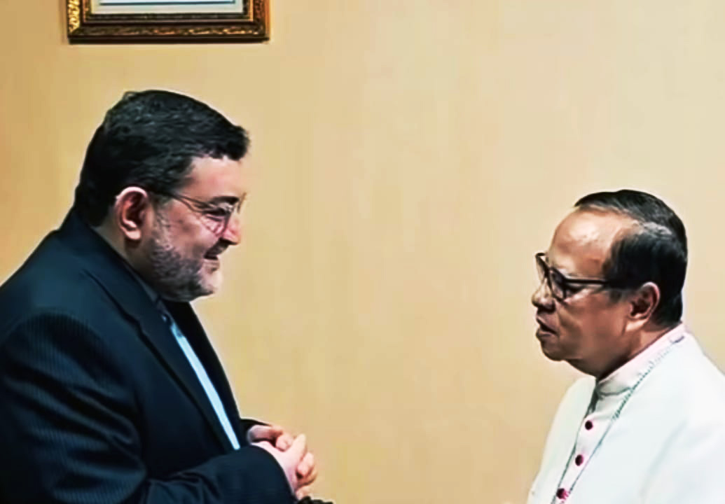 Two men in conversation one in a dark suit with hands clasped the other in white clerical attire with glasses nearby