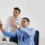 Two business colleagues review something on a computer screen one sits at the desk pointing at the monitor while the other stands beside him looking on
