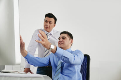 Two business colleagues review something on a computer screen; one sits at the desk pointing at the monitor while the other stands beside him, looking on.