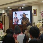 Man speaks at a podium during a formal event addressing an audience with a large screen behind showing another man in batik