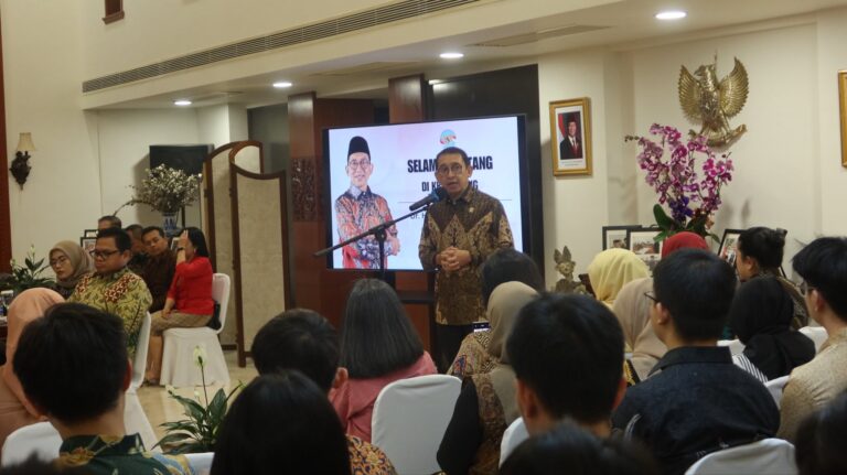Man speaks at a podium during a formal event, addressing an audience with a large screen behind showing another man in batik.