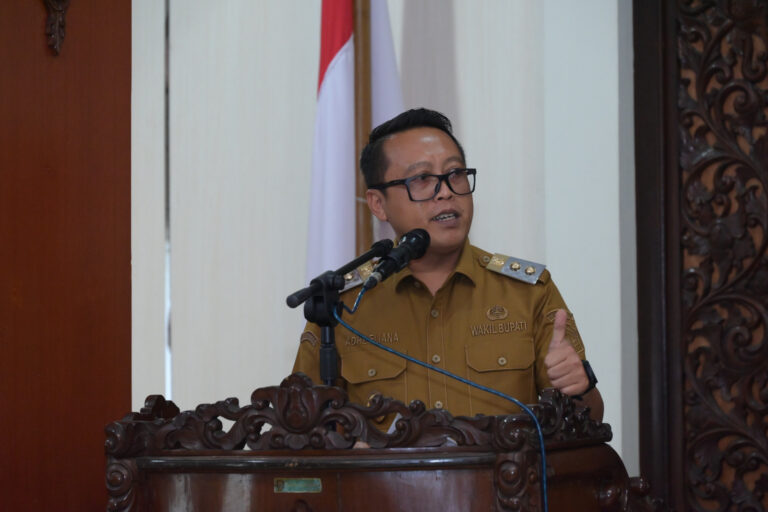 Official in brown uniform speaks at a carved wooden podium with a microphone in front of a flag backdrop.