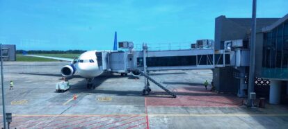 Airplane at gate connected to jet bridge, ground crew around the tarmac under a clear blue sky