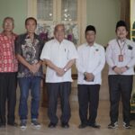 Seven men stand in a row indoors for a formal group portrait wearing batik and white shirts facing the camera with neutral expressions