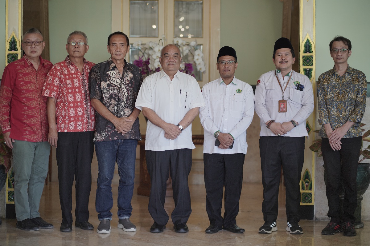 Seven men stand in a row indoors for a formal group portrait wearing batik and white shirts facing the camera with neutral expressions
