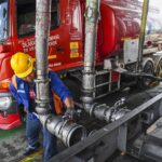 Worker in a yellow hard hat connects large metal hoses to a red tanker truck on an industrial platform far side