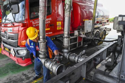 Worker in a yellow hard hat connects large metal hoses to a red tanker truck on an industrial platform far side.