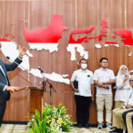 Man in a suit speaks at a podium during a formal event with a wooden backdrop showing a red and white map and an attentive audience behind him and to the side