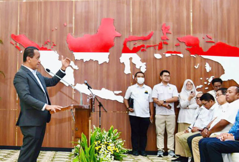Man in a suit speaks at a podium during a formal event, with a wooden backdrop showing a red-and-white map and an attentive audience behind him and to the side.