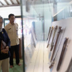 Woman in a headscarf and badge observes a glass display case holding knives in a museum setting