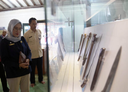 Woman in a headscarf and badge observes a glass display case holding knives, in a museum setting.