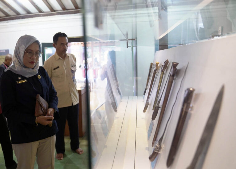 Woman in a headscarf and badge observes a glass display case holding knives, in a museum setting.