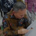 Man in a colorful batik shirt signs a white fabric as several people stand nearby watching the ceremony