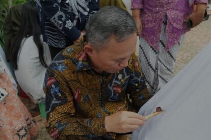 Man in a colorful batik shirt signs a white fabric as several people stand nearby, watching the ceremony.