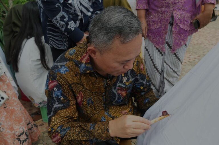 Man in a colorful batik shirt signs a white fabric as several people stand nearby, watching the ceremony.