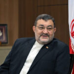 Middle aged man in a dark suit and white shirt sits indoors with a framed picture behind him and the iranian flag to his right