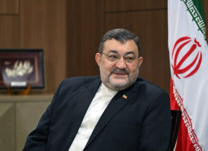 Middle-aged man in a dark suit and white shirt sits indoors with a framed picture behind him and the Iranian flag to his right.
