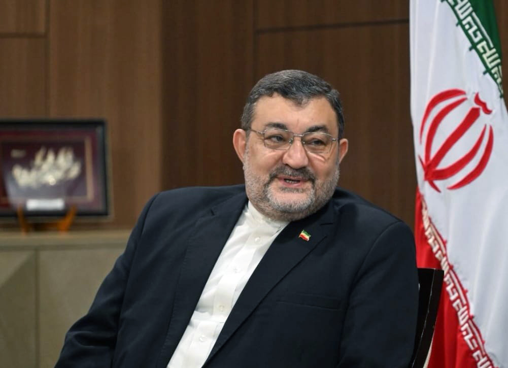 Middle aged man in a dark suit and white shirt sits indoors with a framed picture behind him and the iranian flag to his right