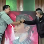 Two men shake hands over a large portrait of a mans face indoors with wooden walls and a pink toned painting in the background