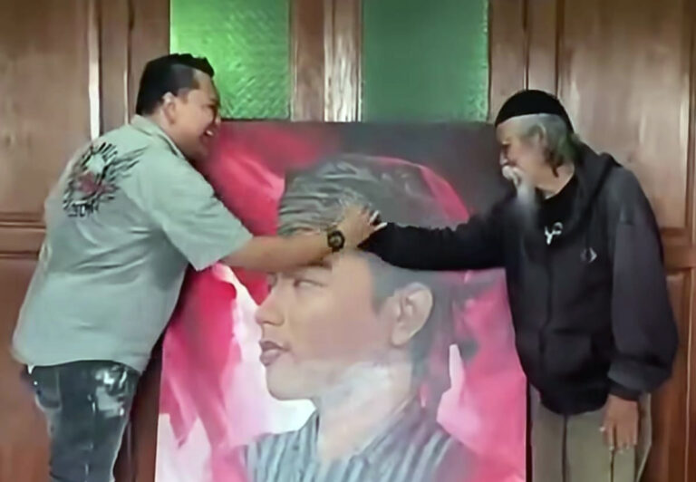 Two men shake hands over a large portrait of a man's face, indoors with wooden walls and a pink-toned painting in the background.