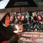 Woman in a black hijab holds a lit candle under an umbrella at a nighttime protest with a crowd and banners in the background