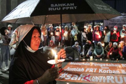 Woman in a black hijab holds a lit candle under an umbrella at a nighttime protest with a crowd and banners in the background