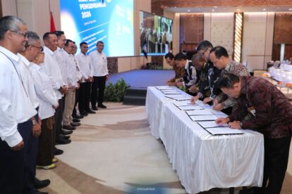 Officials in white shirts stand in a line as delegates sign documents at a long, draped table during a formal signing ceremony.