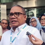 Man in a white shirt and glasses speaks to reporters outside a glass front building a microphone held toward him as others look on
