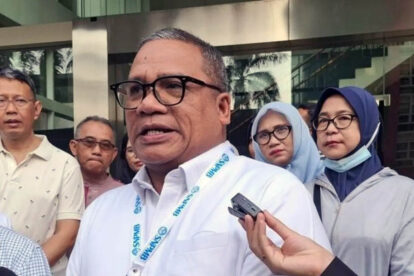 Man in a white shirt and glasses speaks to reporters outside a glass front building a microphone held toward him as others look on