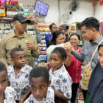 Group of children in matching uniforms in a crowded shop as adults greet them and take photos with smartphones nearby